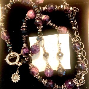 May include: A necklace and earring set featuring large, round, purple beads interspersed with smaller, faceted black beads. The necklace has a silver clasp with a sunflower charm. The earrings have matching purple beads and silver accents. The jewelry is displayed on a black background.
