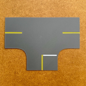May include: A gray, road-shaped play mat with yellow and white lane markings. The mat has a T-intersection design, suitable for toy cars and small vehicles. The mat is made of a durable material.