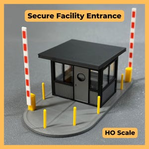 May include: An HO scale model of a secure facility entrance featuring a small black guard booth with a gray door, surrounded by a gray base with yellow bollards and two vertical white and red striped barrier arms. Text reads Secure Facility Entrance.