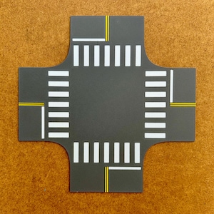 May include: A gray, cross-shaped toy road segment with white pedestrian crosswalks and yellow lane markings. The road has a curved edge on each of the four sides. The toy road is designed for use with toy cars and other vehicles.