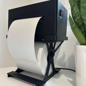 May include: A black label printer with a large roll of white paper. The printer is mounted on a black stand. A green plant in a white pot is in the background.