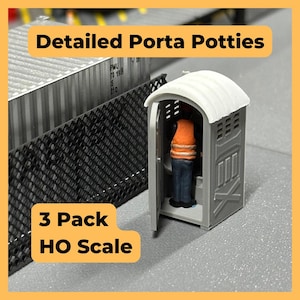 May include: A miniature gray porta potty with an open door, featuring a figure wearing an orange vest and blue pants. The text "Detailed Porta Potties" and "3 Pack HO Scale" are displayed on the image. A model train accessory.