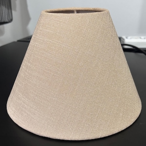 May include: A beige lampshade with a conical shape. The lampshade is made from a textured fabric and has a neutral colour. The lampshade is designed to fit on a lamp base and diffuse light.