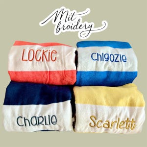 May include: Four personalised beach towels with embroidered names. Each towel has a white stripe with a name embroidered in a contrasting colour. The towels are in shades of red, blue, yellow, and navy.