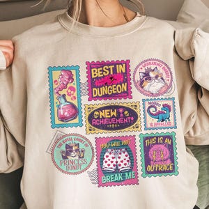 May include: Beige sweatshirt with colorful stamp-like graphics. The stamps feature various designs, including a cat, a potion, and text that reads "BEST IN DUNGEON", "NEW ACHIEVEMENT", and "THIS IS AN OUTRACE".