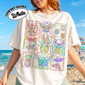 May include: A white short-sleeve t-shirt featuring a colorful graphic print of various insects, including beetles, moths, and a praying mantis, arranged in a grid-like pattern with decorative borders and small star accents throughout the design.