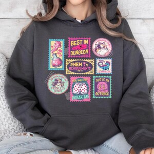 May include: Dark gray hoodie featuring a collection of colorful stamp-like graphics. The stamps display various designs, including a potion bottle, a cat, a dinosaur, and text that reads "BEST IN DUNGEON", "NEW ACHIEVEMENT", "PRINCESS DONUT", and "BREAK ME".