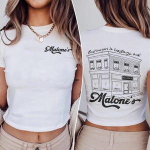 May include: A white short-sleeved t-shirt featuring the black text Malone's on the front chest. The back displays a black line drawing of a building with the text Best burgers in hastings. ma! and the word Malone's in a large script font.