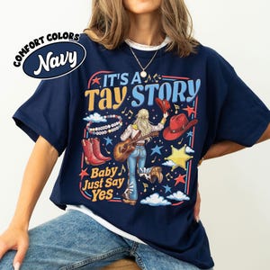 Pu&ograve; includere: Una t-shirt blu navy con una stampa grafica con il testo IT'S A TAY STORY e Baby Just Say Yes. Il design include una persona con lunghi capelli biondi che suona la chitarra, stivali da cowboy rossi, un cappello rosso, un braccialetto e stelle.