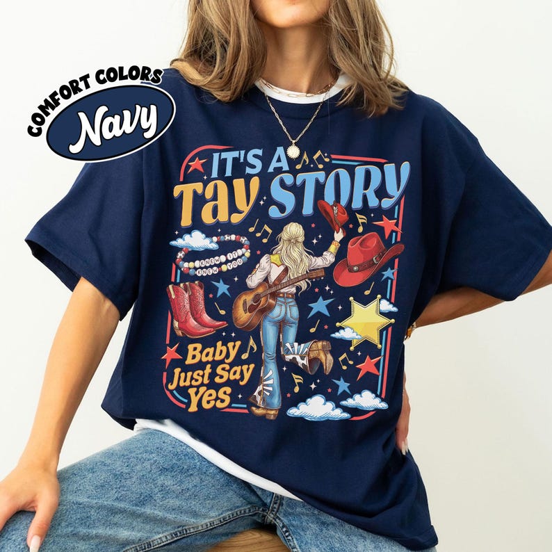 Pu&ograve; includere: Una t-shirt blu navy con una stampa grafica con il testo IT'S A TAY STORY e Baby Just Say Yes. Il design include una persona con lunghi capelli biondi che suona la chitarra, stivali da cowboy rossi, un cappello rosso, un braccialetto e stelle.