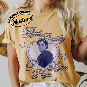 May include: Mustard-colored t-shirt with the text "But Daddy I Love Him" in a cursive font. The shirt features a heart-shaped graphic with a portrait of a man. The words "Comfort Colors" are above the word "Mustard".
