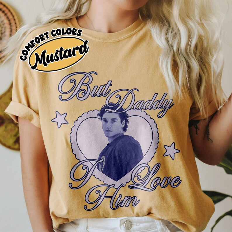 May include: Mustard-colored t-shirt with the text "But Daddy I Love Him" in a cursive font. The shirt features a heart-shaped graphic with a portrait of a man. The words "Comfort Colors" are above the word "Mustard".