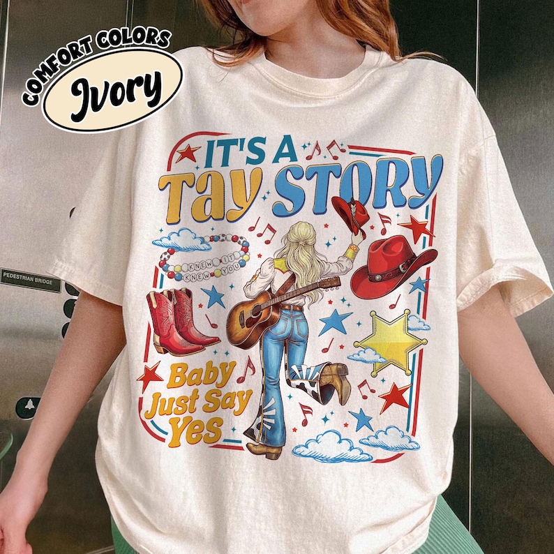 Pu&ograve; includere: Una t-shirt color avorio con una stampa grafica che riporta il testo IT'S A TAY STORY e Baby Just Say Yes. Il design raffigura una persona con capelli biondi che suona la chitarra, stivali da cowboy rossi, un cappello rosso, stelle e nuvole.