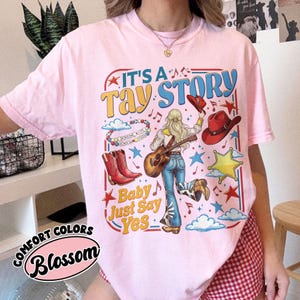 Pu&ograve; includere: Una t-shirt rosa chiaro con una stampa grafica che riporta il testo IT'S A TAY STORY e Baby Just Say Yes. Il design include una persona con capelli biondi che tiene una chitarra, stivali da cowboy rossi, un cappello rosso, stelle e nuvole.