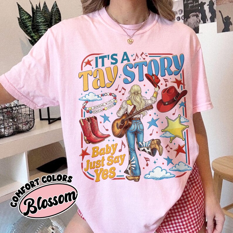 Pu&ograve; includere: Una t-shirt rosa chiaro con una stampa grafica che riporta il testo IT'S A TAY STORY e Baby Just Say Yes. Il design include una persona con capelli biondi che tiene una chitarra, stivali da cowboy rossi, un cappello rosso, stelle e nuvole.