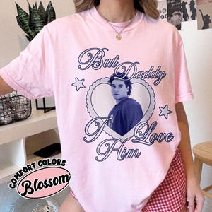 May include: A light pink t-shirt with the text "But Daddy I Love Him" in a cursive font, surrounding a heart-shaped image of a person. The shirt also features the words "Comfort Colors" and "Blossom".