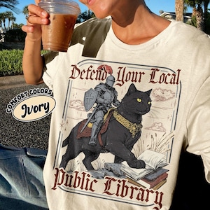 May include: Ivory-colored t-shirt with a graphic of a knight riding a black cat, with the text "Defend Your Local Public Library." The knight holds a shield and sword. The shirt also has the words "Comfort Colors" and "Ivory" printed on it.