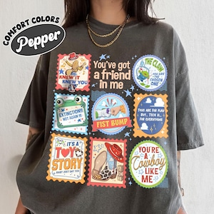 Puede incluir: Una camiseta gris carb&oacute;n con un collage de sellos gr&aacute;ficos coloridos y texto. Los dise&ntilde;os incluyen un vaquero, una m&aacute;quina de garra, un choque de pu&ntilde;os, una rana, una bota y frases como You've got a friend in me y The Claw.