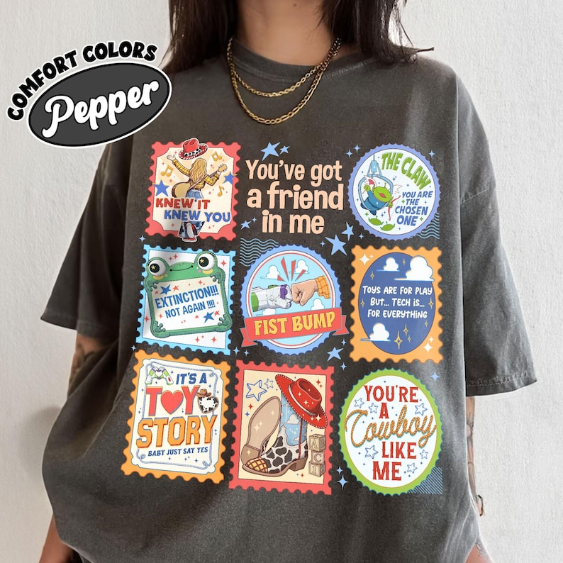 Puede incluir: Una camiseta gris carb&oacute;n con un collage de sellos gr&aacute;ficos coloridos y texto. Los dise&ntilde;os incluyen un vaquero, una m&aacute;quina de garra, un choque de pu&ntilde;os, una rana, una bota y frases como You've got a friend in me y The Claw.