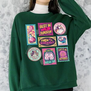 May include: A dark green sweatshirt featuring a collection of colorful postage stamp-style graphics. The stamps display various images and phrases, including "BEST IN DUNGEON," "NEW ACHIEVEMENT," and "BREAK ME." The sweatshirt has a crew neck and long sleeves.