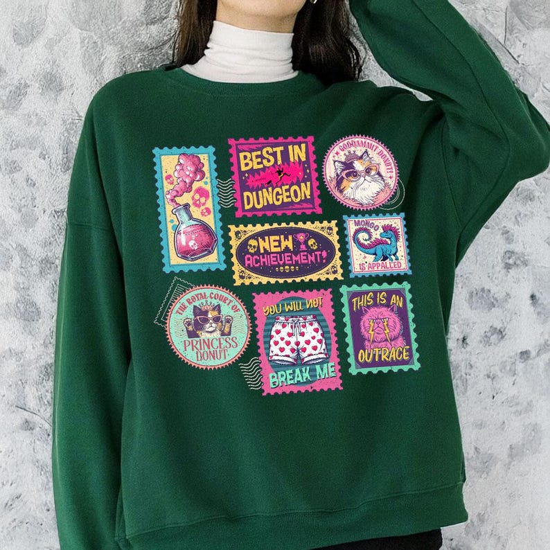 May include: A dark green sweatshirt featuring a collection of colorful postage stamp-style graphics. The stamps display various images and phrases, including "BEST IN DUNGEON," "NEW ACHIEVEMENT," and "BREAK ME." The sweatshirt has a crew neck and long sleeves.