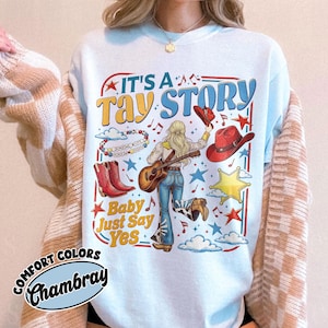 Pu&ograve; includere: Una t-shirt azzurra con una stampa grafica con il testo IT'S A TAY STORY e Baby Just Say Yes. Il design include una persona con capelli biondi che suona la chitarra, stivali da cowboy rossi, un cappello rosso, una stella, nuvole e note musicali.