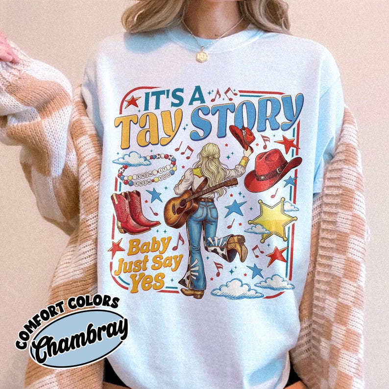 Pu&ograve; includere: Una t-shirt azzurra con una stampa grafica con il testo IT'S A TAY STORY e Baby Just Say Yes. Il design include una persona con capelli biondi che suona la chitarra, stivali da cowboy rossi, un cappello rosso, una stella, nuvole e note musicali.