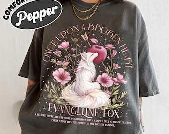 May include: A charcoal gray t-shirt featuring a graphic of a white fox surrounded by pink flowers and a red apple pierced by an arrow. The text reads "ONCE UPON A BROKEN HEART", "EVANGELINE FOX", and "I BELIEVE THERE ARE FAR MORE POSSIBILITIES THAN HAPPILY EVER AFTER OR TRAGEDY EVERY STORY HAS THE POTENTIAL FOR INFINITE ENDINGS."