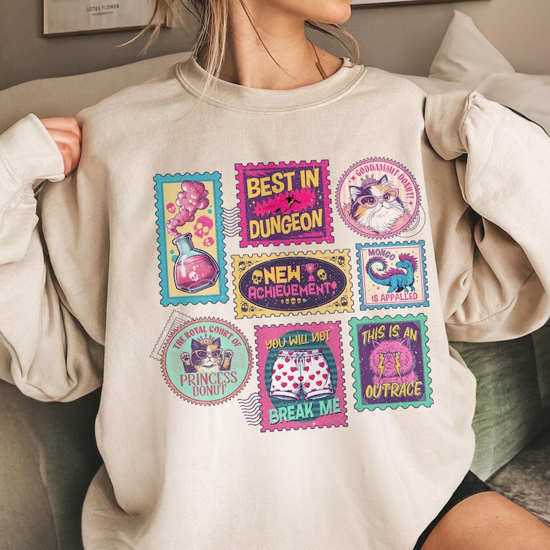 May include: Beige sweatshirt with colorful stamp-like graphics. The stamps feature various designs, including a cat, a potion, and text that reads "BEST IN DUNGEON", "NEW ACHIEVEMENT", and "THIS IS AN OUTRACE".