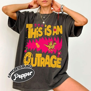 May include: Dark gray t-shirt with a distressed yellow and pink graphic that reads "This is an Outrage." The design includes a dinosaur, pineapple, and donut. The shirt also has the text "Comfort Colors Pepper."