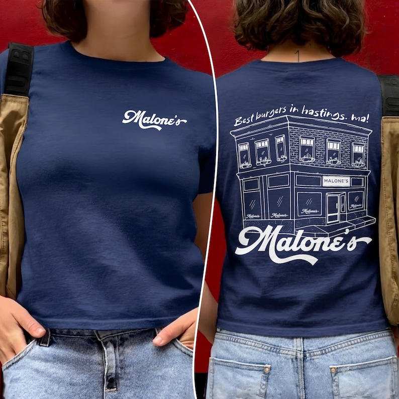 May include: A navy blue t-shirt featuring a white script logo on the front chest that reads Malone's. The back displays a white line drawing of a building with the text Best burgers in hastings. ma! and the script logo Malone's below it.