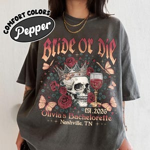 May include: A charcoal gray t-shirt featuring a graphic with the text "Bride or Die", "Est. 2026", "Olivia's Bachelorette", and "Nashville, TN". The design includes a skull wearing a crown, red roses, butterflies, and a glass of red wine.