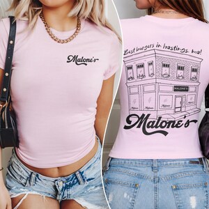 May include: A light pink short-sleeve t-shirt featuring the black text Malone's on the front chest. The back displays a black line-art illustration of a building with the text Best burgers in hastings. ma! and the script logo Malone's.