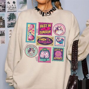 May include: A cream-colored sweatshirt featuring colorful stamp-like graphics. The stamps display various images and phrases, including "Best in Dungeon," "Princess Donut," and "This is an Outrace." The design incorporates pink, blue, and yellow hues.