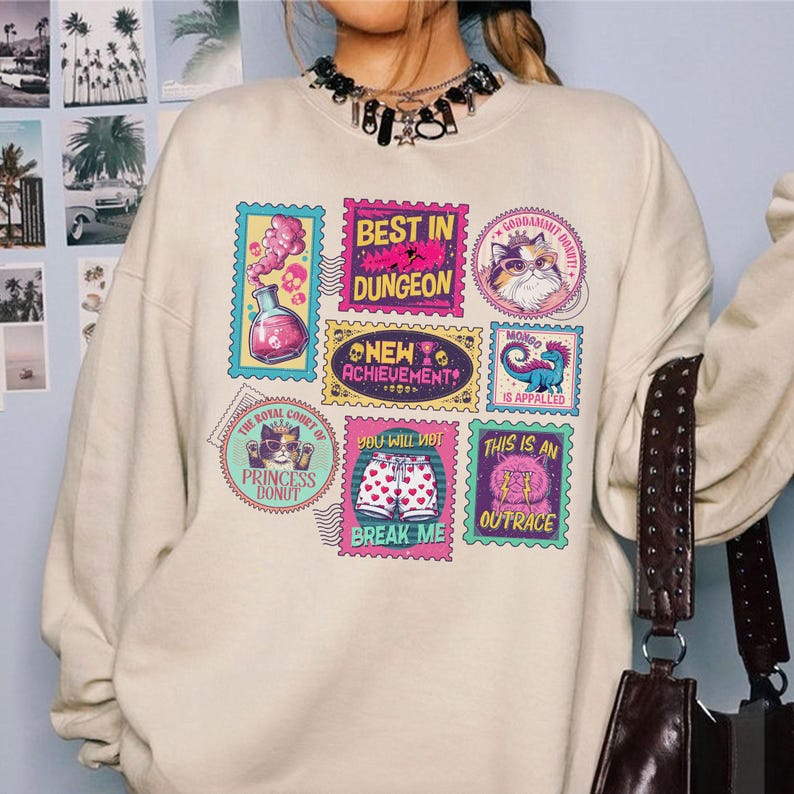 May include: A cream-colored sweatshirt featuring colorful stamp-like graphics. The stamps display various images and phrases, including "Best in Dungeon," "Princess Donut," and "This is an Outrace." The design incorporates pink, blue, and yellow hues.
