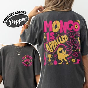 May include: A charcoal gray short-sleeve t-shirt featuring a large graphic on the back with the text "MONGO IS APPALLED" in pink and yellow, accompanied by a dinosaur illustration, a trophy, and a bomb. A smaller version is on the front left chest.