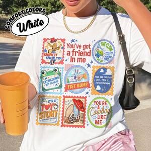 Puede incluir: Una camiseta blanca con una cuadr&iacute;cula de sellos gr&aacute;ficos coloridos con varias ilustraciones de dibujos animados y texto que incluye "You've got a friend in me," "The Claw," "Fist Bump," "It's a Toy Story," y "You're a Cowboy Like Me".