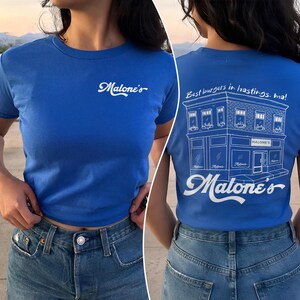 May include: A bright blue t-shirt featuring a white script logo on the front that reads Malone's. The back displays a white line drawing of a building with the text Best burgers in hastings. ma! and the script logo Malone's at the bottom.