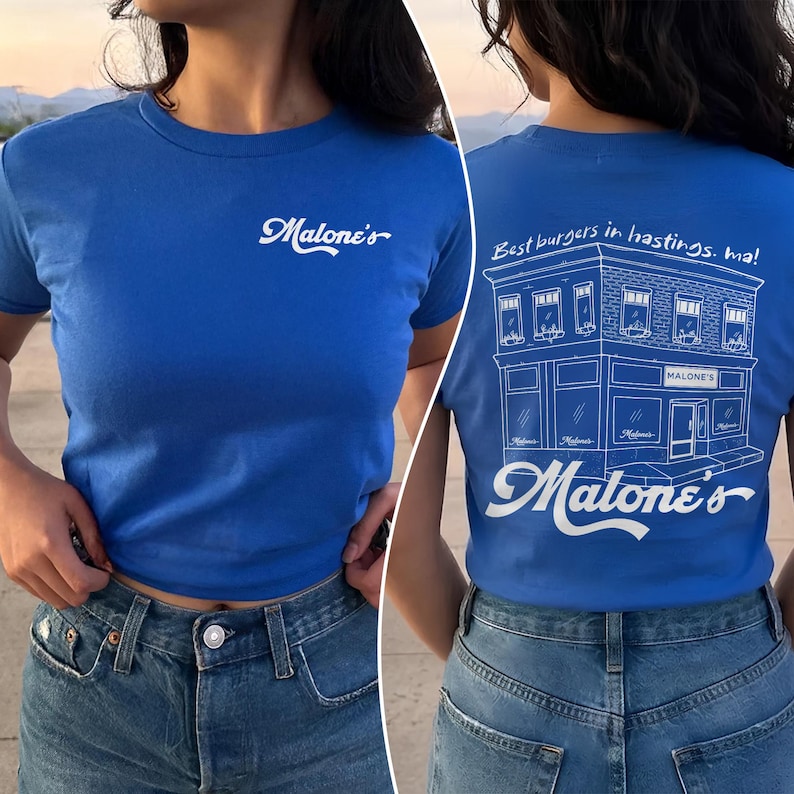May include: A bright blue t-shirt featuring a white script logo on the front that reads Malone's. The back displays a white line drawing of a building with the text Best burgers in hastings. ma! and the script logo Malone's at the bottom.