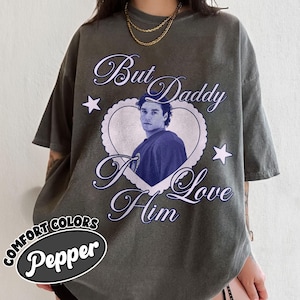 May include: A dark grey t-shirt with the text "But Daddy I Love Him" in a cursive font, surrounding a heart-shaped image of a person. The shirt also features two white stars and the words "Comfort Colors Pepper" in an oval.
