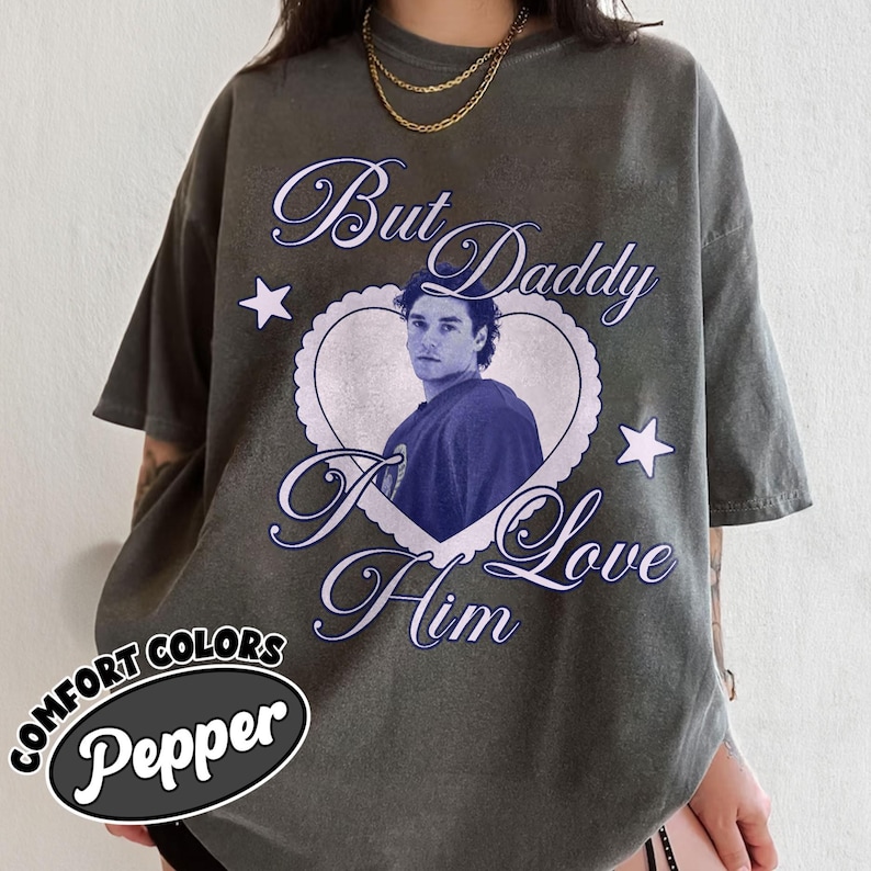 May include: A dark grey t-shirt with the text "But Daddy I Love Him" in a cursive font, surrounding a heart-shaped image of a person. The shirt also features two white stars and the words "Comfort Colors Pepper" in an oval.