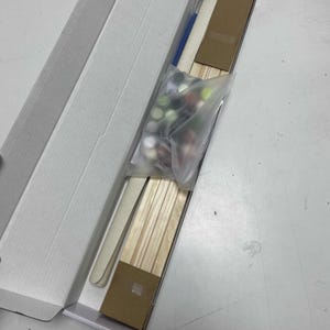 May include: An open white cardboard box containing a paint-by-numbers kit. The kit includes a small blue paintbrush, wooden sticks, a plastic bag of paint pots, and a cardboard insert.