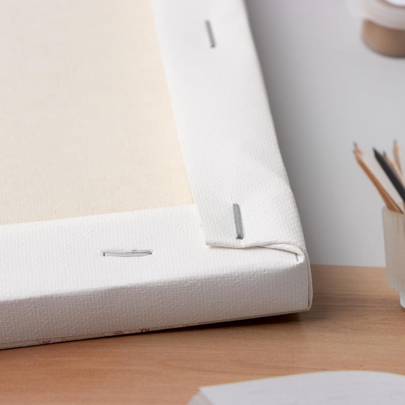 May include: Close-up of a white canvas stretched over a wooden frame. The canvas is secured with staples along the edges. The canvas is a light beige color, and the frame is white.