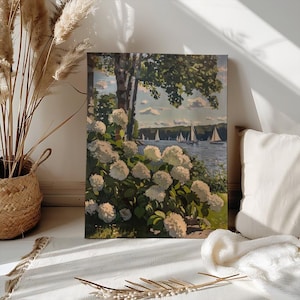 May include: A framed landscape painting depicts a serene lake scene with sailboats, lush green trees, and white hydrangea bushes in the foreground. The artwork is displayed indoors, leaning against a wall, with natural light illuminating the scene.