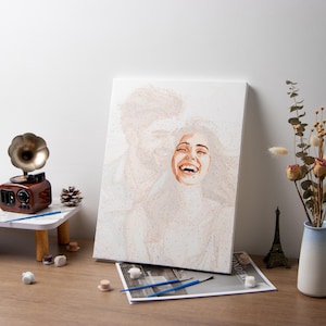 May include: A paint-by-numbers canvas depicts a woman laughing with her head tilted back. The painting is mostly shades of white and beige. A small wooden record player, paintbrushes, and other art supplies are on a table.