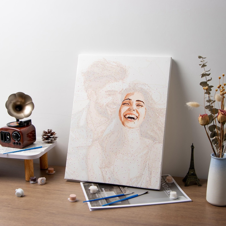 May include: A paint-by-numbers canvas depicts a woman laughing with her head tilted back. The painting is mostly shades of white and beige. A small wooden record player, paintbrushes, and other art supplies are on a table.