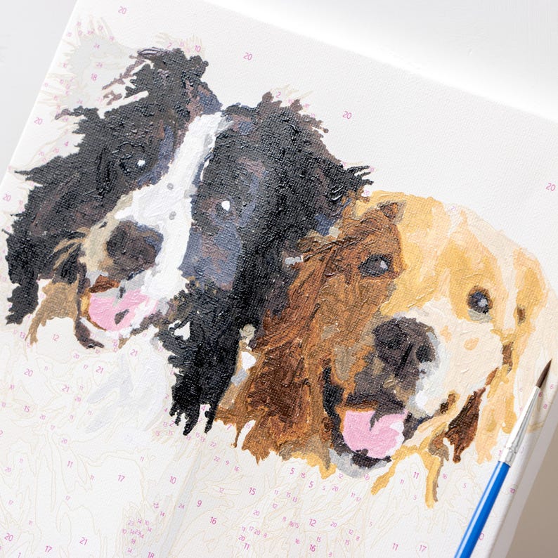 May include: A paint-by-numbers canvas featuring three dog portraits. The dogs are painted in shades of black, brown, and gold, with pink tongues. A blue paintbrush is visible on the right side of the canvas. The canvas has numbers and paint sections.