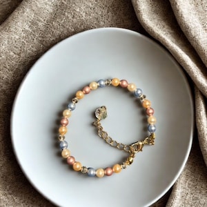 Puede incluir: Una pulsera de cuentas con un cierre y una cadena extensora dorados. La pulsera presenta una mezcla de cuentas de colores pastel en tonos rosa, amarillo y azul. La pulsera se muestra en un plato azul claro.
