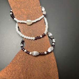 Könnte beinhalten: Drei Armbänder mit Perlen auf einem Holzständer. Die Armbänder haben weiße und lila Perlen mit silberfarbenen, gerippten Akzentperlen. Ein Armband hat eine herzförmige Perle.