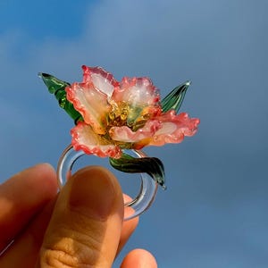 May include: A clear glass ring with a flower design. The flower has pink and yellow petals and green leaves. The ring is held against a blue sky. The ring is a piece of jewellery.